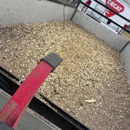 Magiczny Trawnik - Przyczepa wypełniona zrębkami drzewnymi, widoczny fragment czerwonego narzędzia i biały samochód dostawczy Decat w tle. Transport drewna.