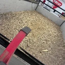 Przyczepa wypełniona zrębkami drzewnymi, widoczny fragment czerwonego narzędzia i biały samochód dostawczy Decat w tle. Transport drewna.