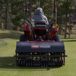 Magiczny Trawnik - Aeracja trawnika: Traktor Toro ProCore z operatorem na zielonym polu golfowym, w tle drzewa i budynki. Widok z tyłu, skupiony na maszynie i efekcie aeracji.