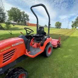 Czerwony traktor Kubota BX2350 na zielonej trawie, obok świeżo skoszony pas. W tle drzewa i błękitne niebo z chmurami. Widok z boku, traktor w ruchu.