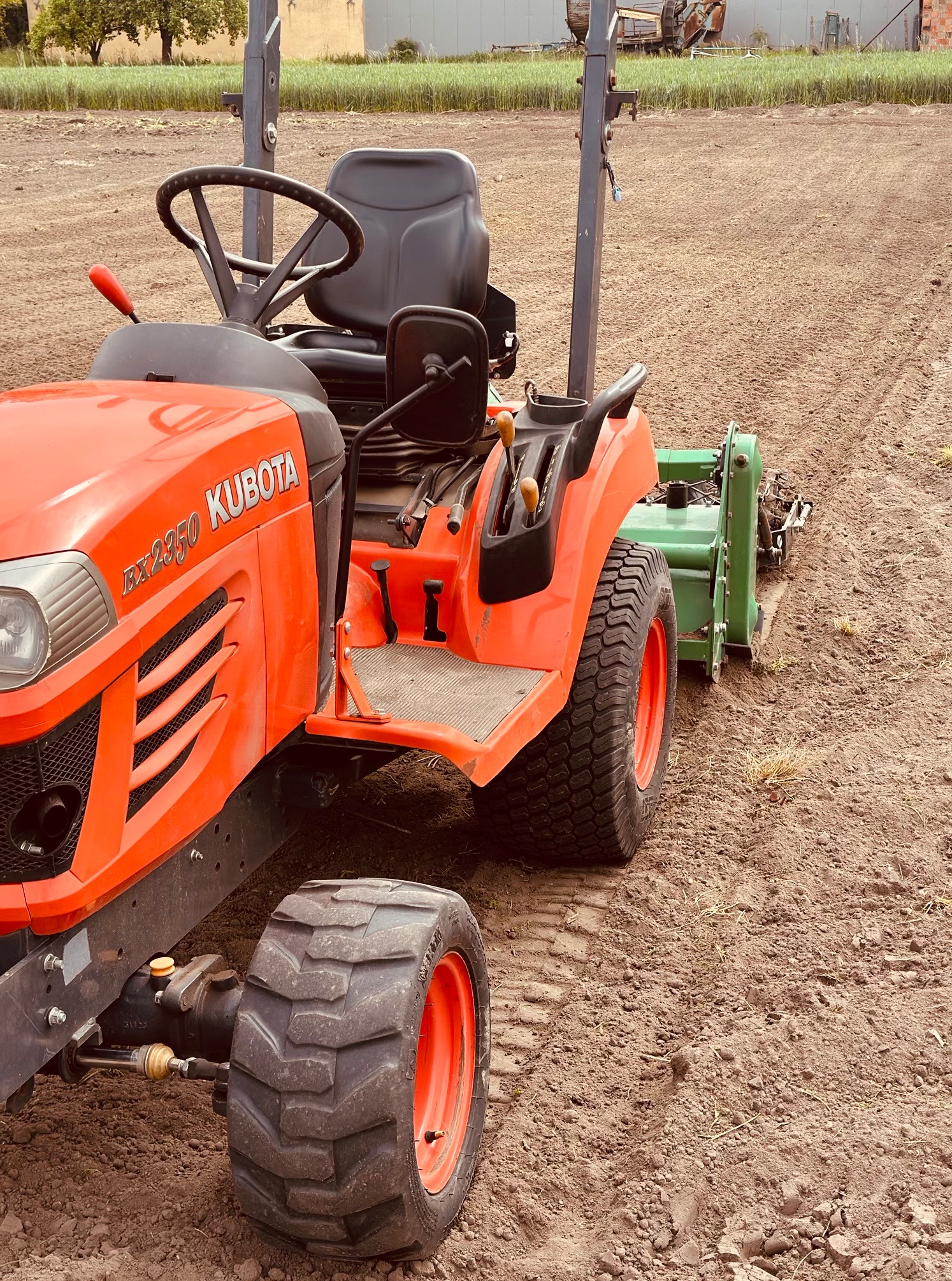 Pomarańczowy traktor Kubota RX2350 na polu, przygotowujący ziemię pod uprawę. Widoczne ślady opon i świeżo spulchniona gleba. Sprzęt rolniczy w trakcie pracy polowej.