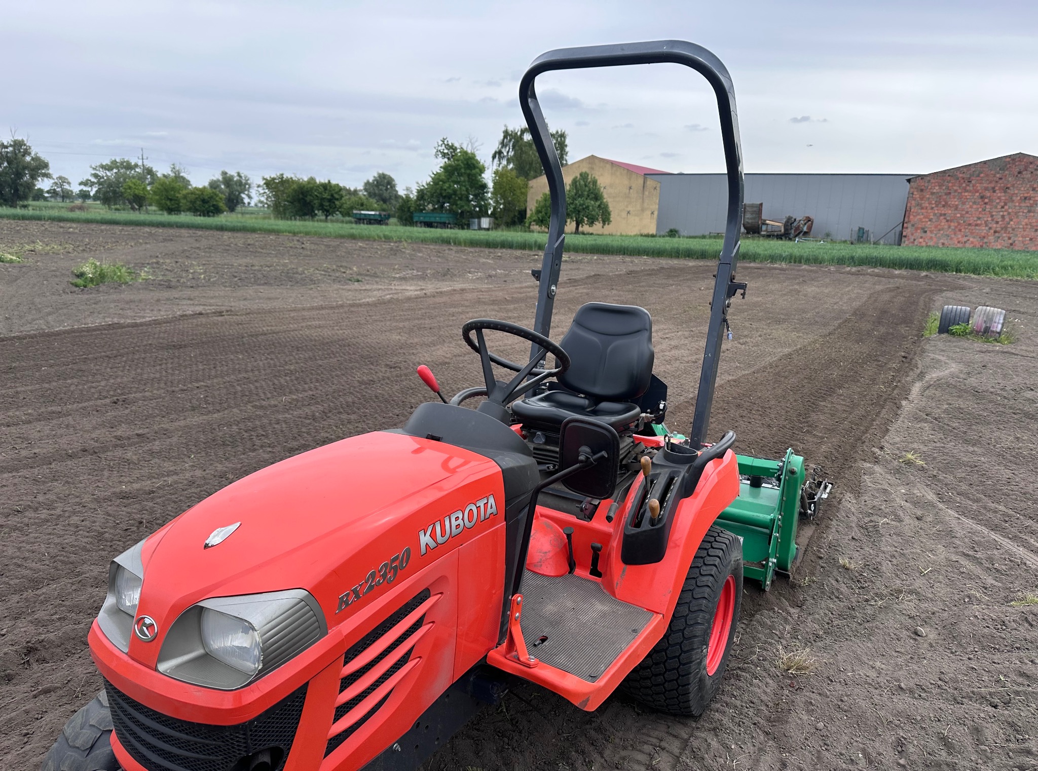 Czerwony traktor Kubota RX2350 z glebogryzarką na polu uprawnym, przygotowanym do siewu. Widoczne budynki gospodarcze w tle.