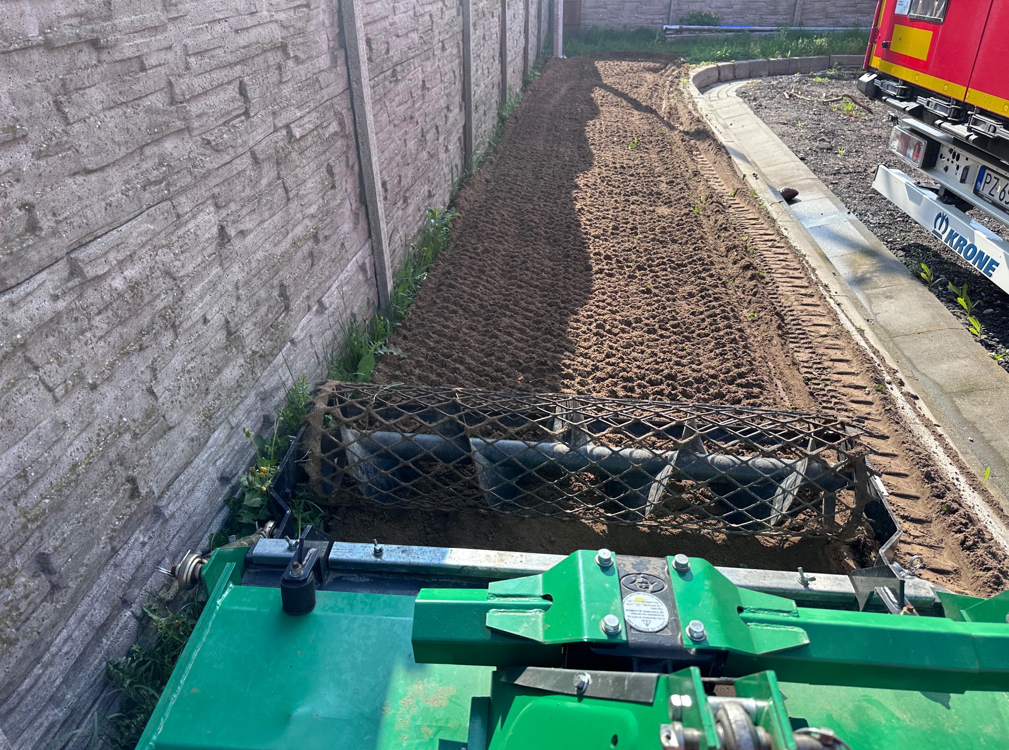 Przygotowanie gleby pod trawnik za pomocą glebogryzarki separacyjnej, widok z perspektywy operatora. W tle betonowy płot i fragment czerwonego samochodu.