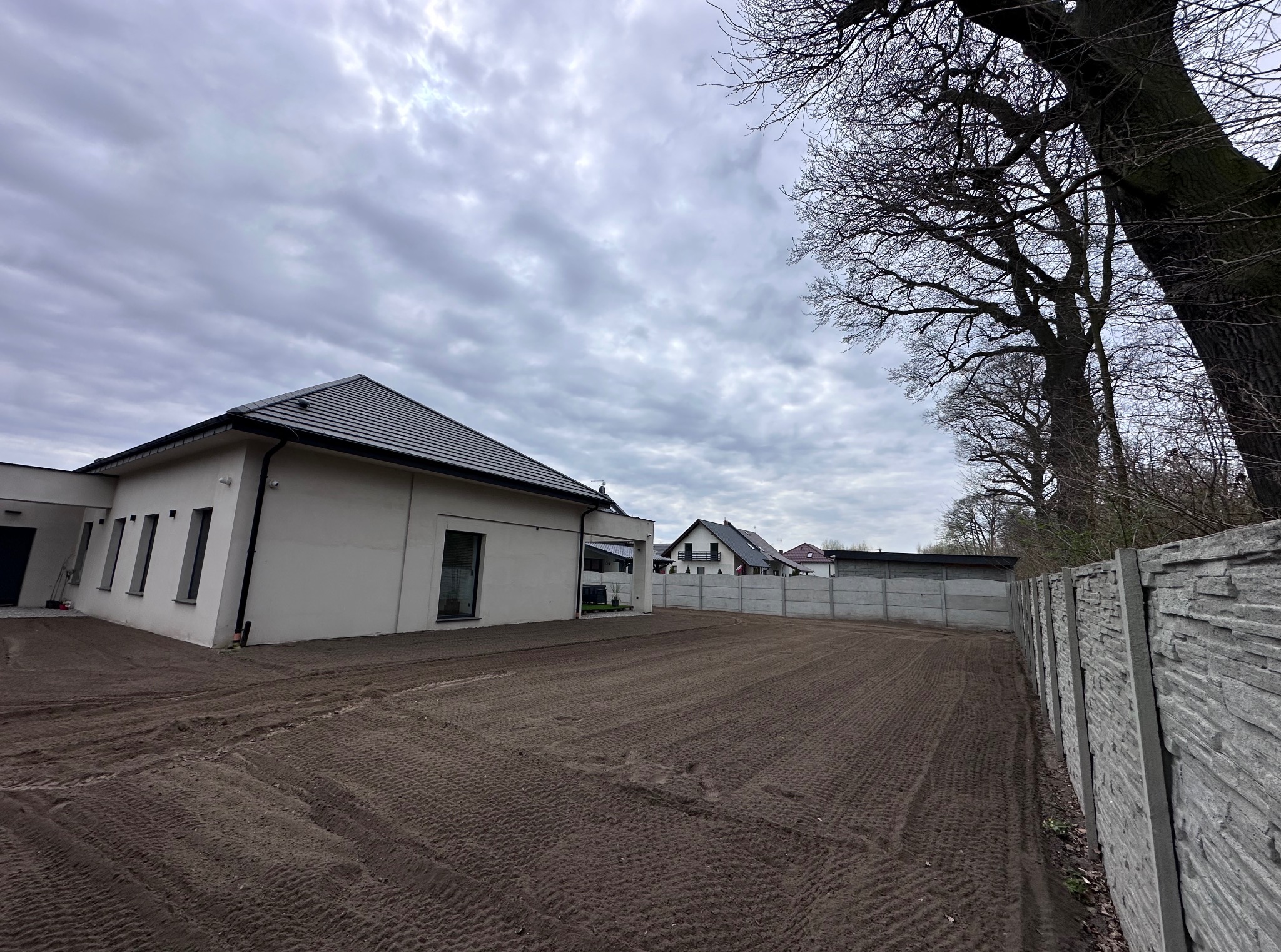 Wyrównany teren pod trawnik przy nowoczesnym domu z szarą dachówką, ogrodzony betonowym płotem. Widoczne ślady po maszynach. Pochmurne niebo w tle.