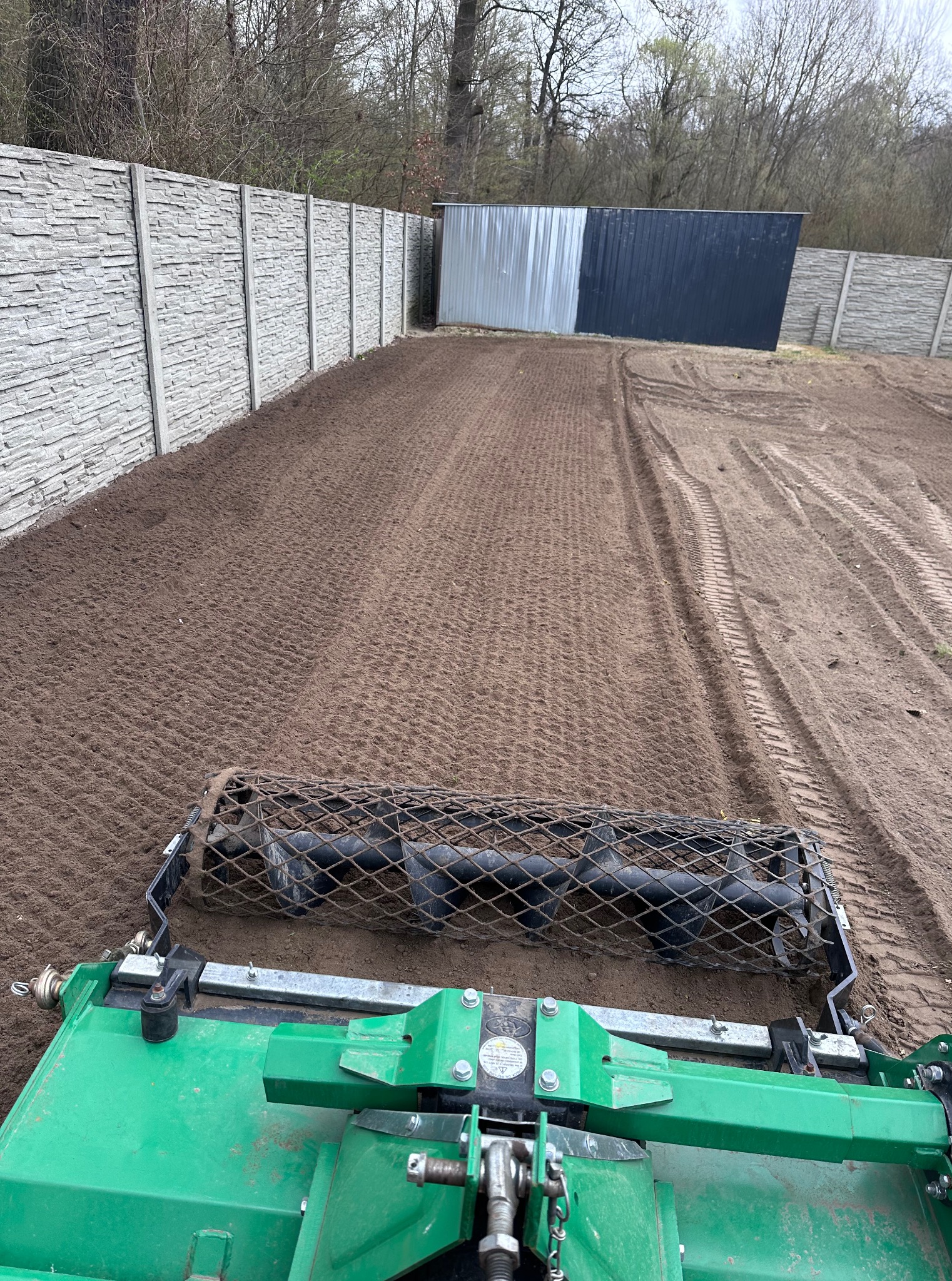 Przygotowanie terenu pod trawnik: walcowanie gleby walcem kolczatkowym. Widoczne ślady opon i wyrównana powierzchnia ziemi w ogrodzie, ogrodzenie betonowe w tle.