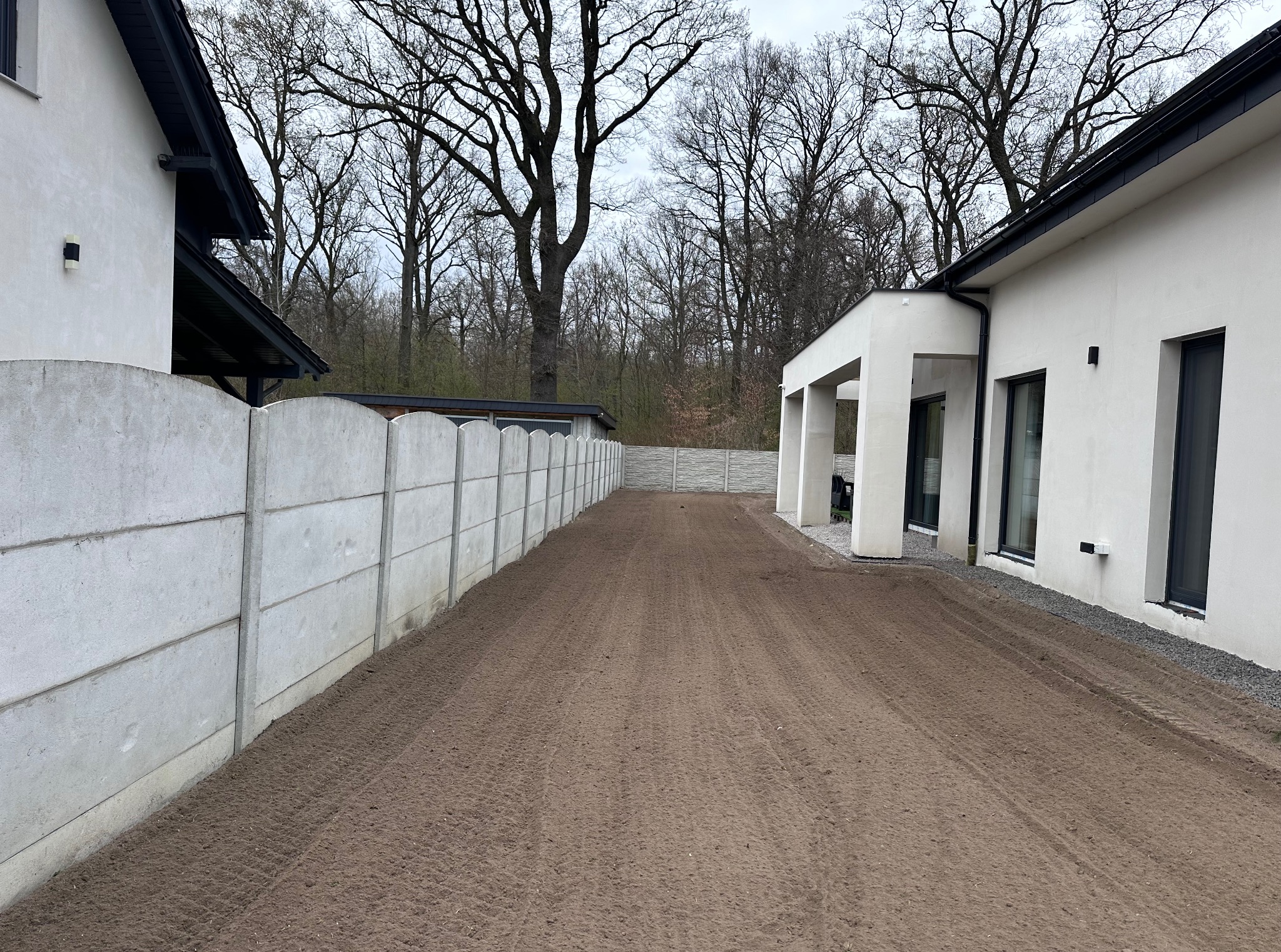 Przygotowany teren pod trawnik, wyrównana ziemia obok nowoczesnego domu z betonowym ogrodzeniem. Widoczne ślady po maszynach.