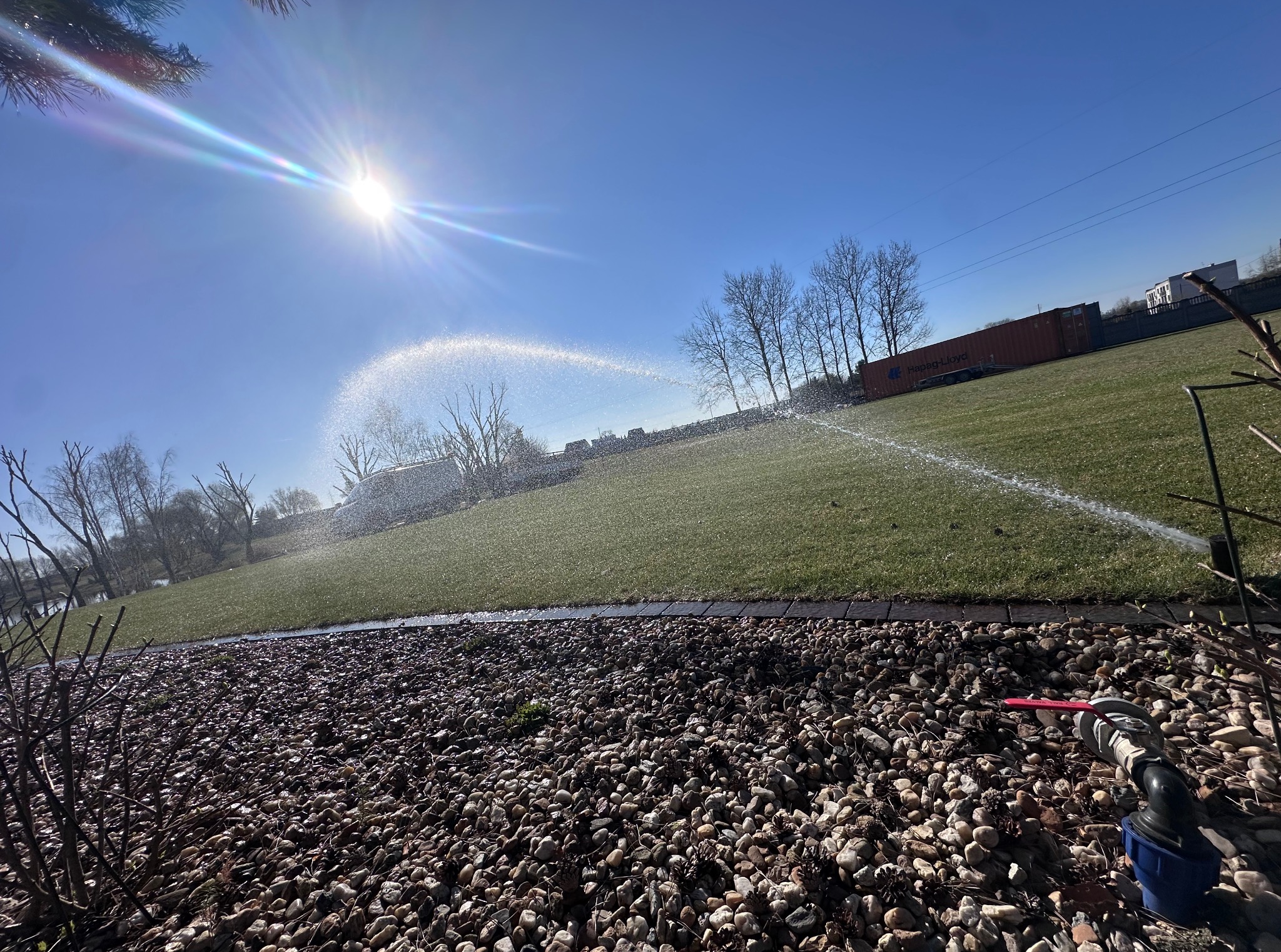 Nawadnianie trawnika zraszaczy, w tle kontener transportowy. Widok na zadbany ogród w słoneczny dzień, z bliska kamienie i zawór.