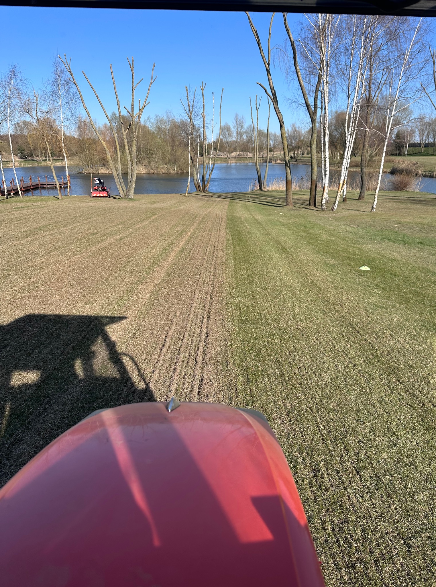 Widok z traktora na świeżo skoszoną trawę w parku z jeziorem i drzewami. W oddali widoczny koszący trawnik. Słoneczny dzień.