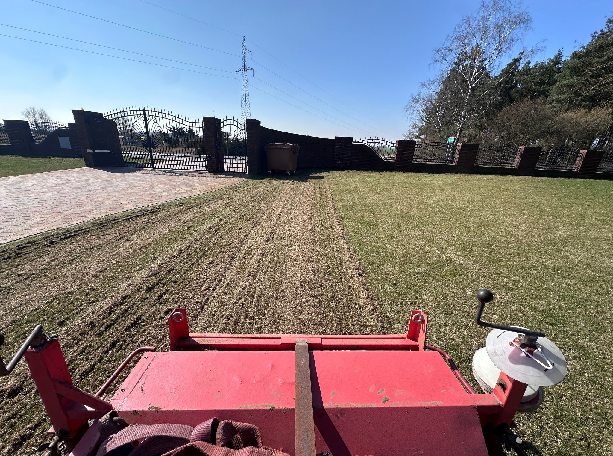 Aeracja trawnika z perspektywy operatora maszyny. Widoczne ślady po aeracji, brama w tle i słup wysokiego napięcia. Słoneczny dzień.