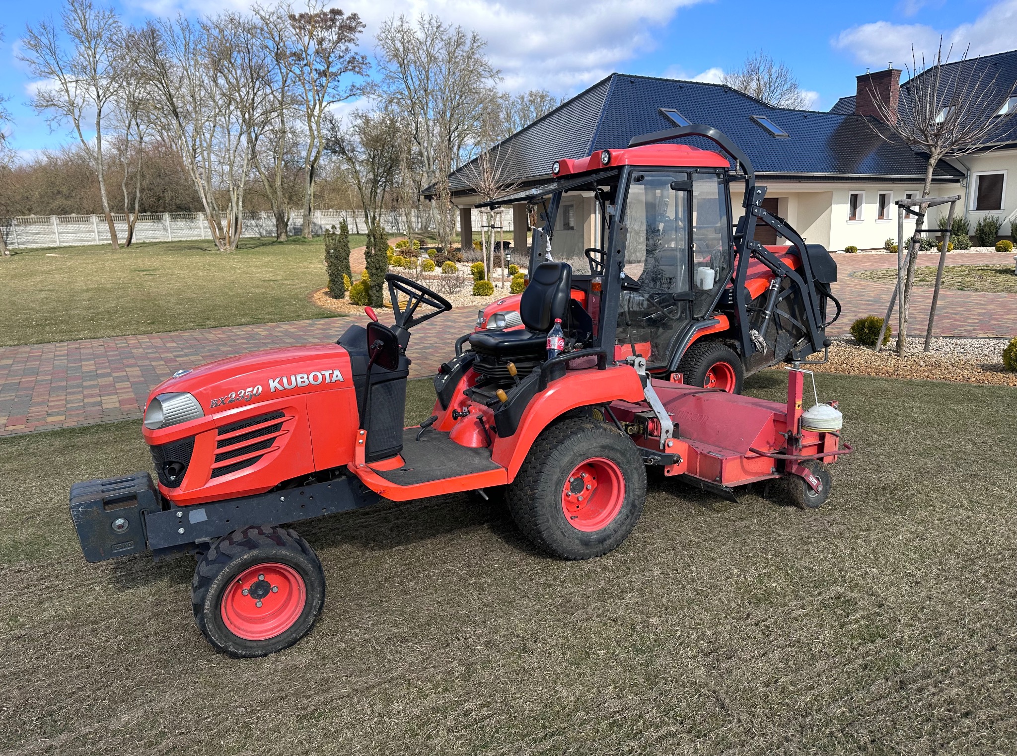 Czerwony traktor Kubota BX2350 z kosiarką na trawniku przed domem. Widoczny drugi traktor w tle, drzewa bez liści i błękitne niebo.