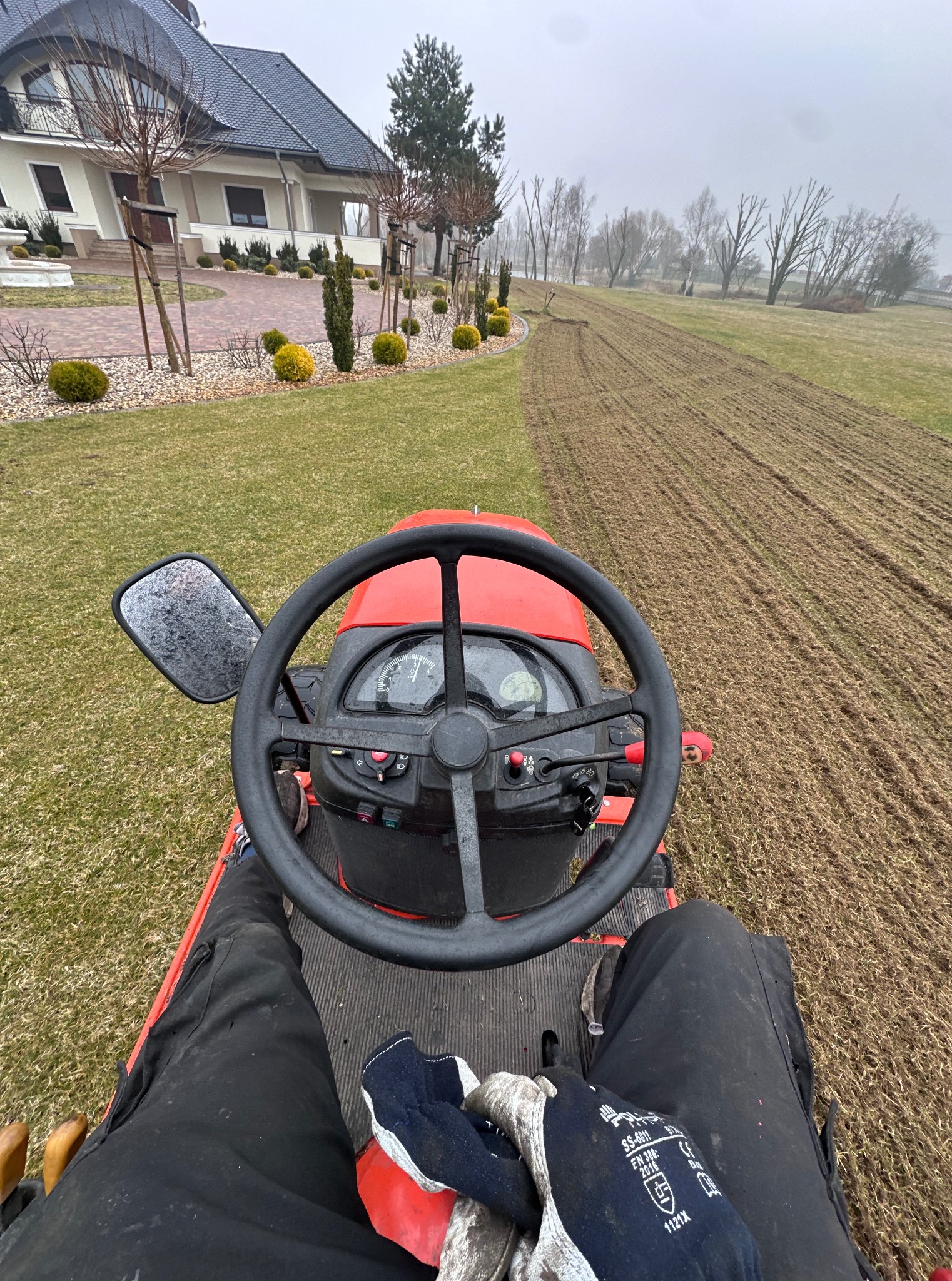 Czerwony traktor na zielonym trawniku, widok z perspektywy operatora podczas wykonywania prac ziemnych na posesji z domem w tle.