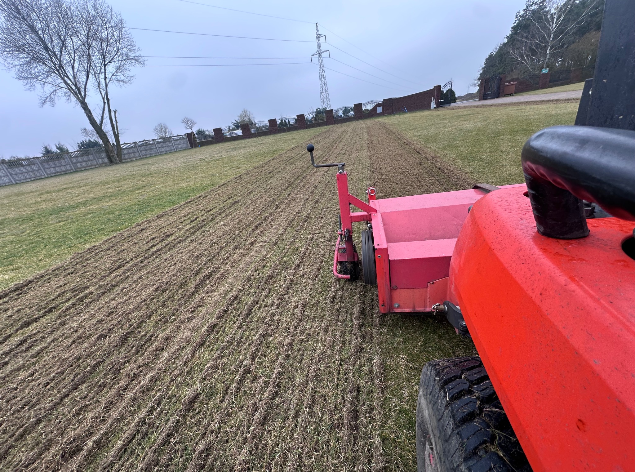 Czerwony traktor z broną talerzową na polu, przygotowujący ziemię pod zasiew. Widok z perspektywy operatora, w tle drzewa i słupy wysokiego napięcia.