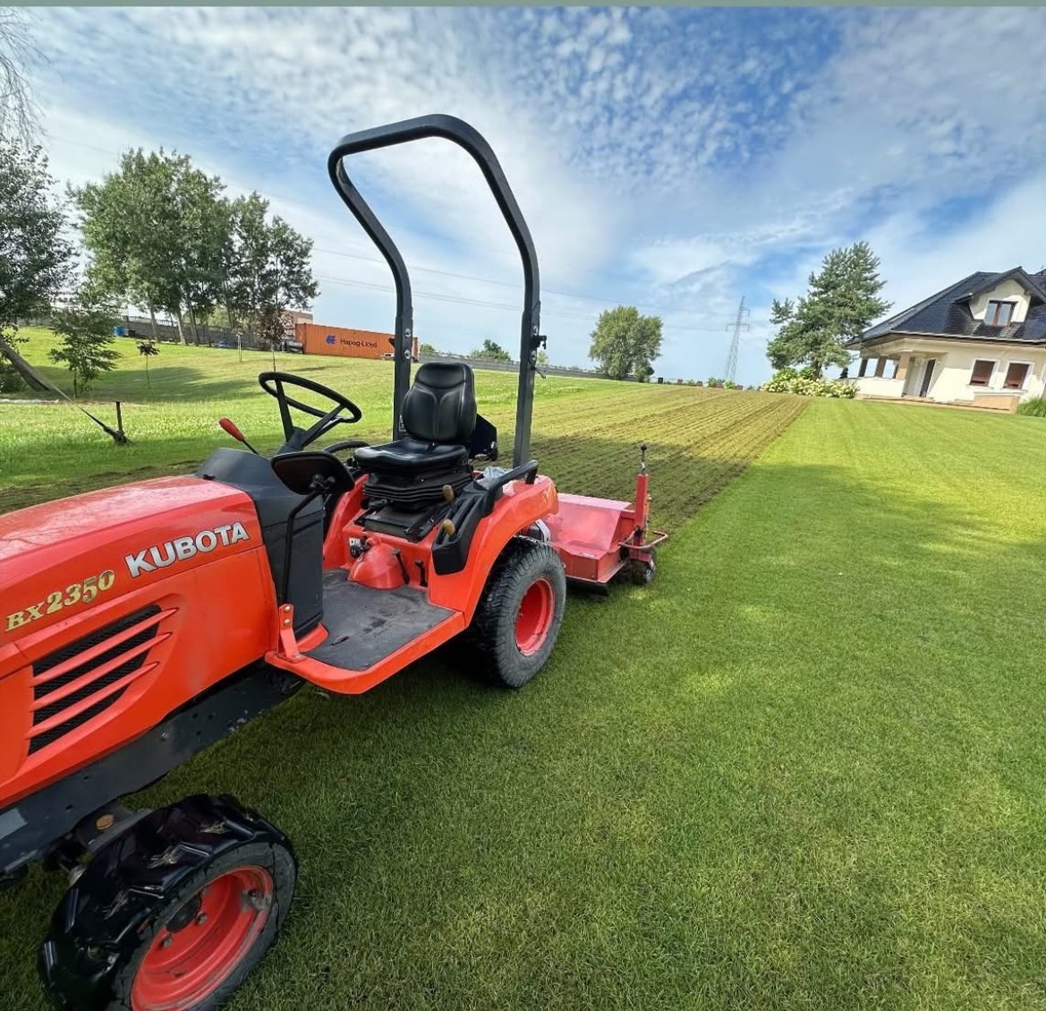 Czerwony traktor Kubota BX2350 na zielonym trawniku po aeracji, z domem w tle i błękitnym niebem. Widok z boku, słoneczny dzień.