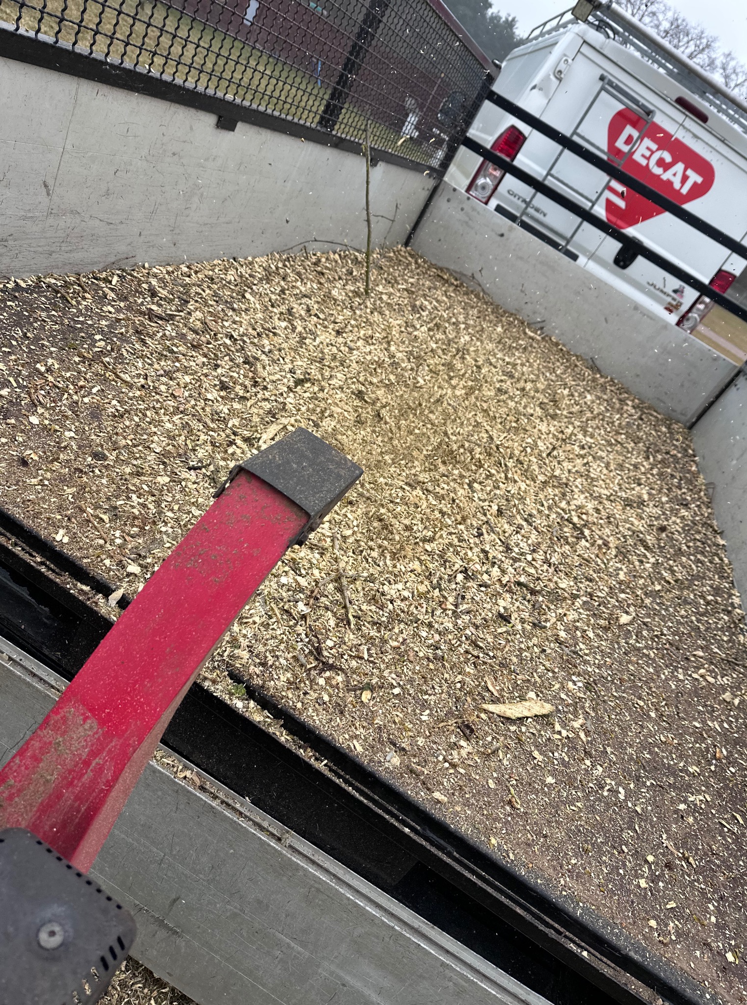 Przyczepa wypełniona zrębkami drzewnymi, widoczny fragment czerwonego narzędzia i biały samochód dostawczy Decat w tle. Transport drewna.