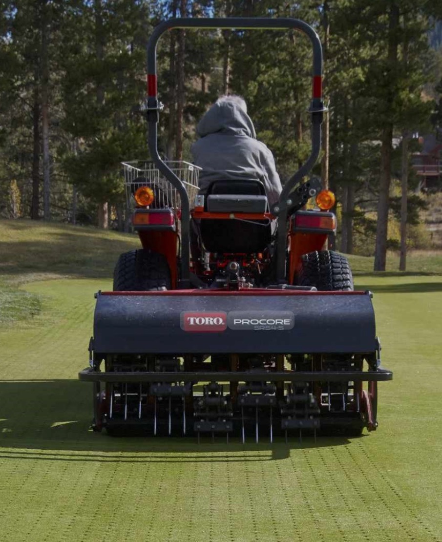 Aeracja trawnika: Traktor Toro ProCore z operatorem na zielonym polu golfowym, w tle drzewa i budynki. Widok z tyłu, skupiony na maszynie i efekcie aeracji.