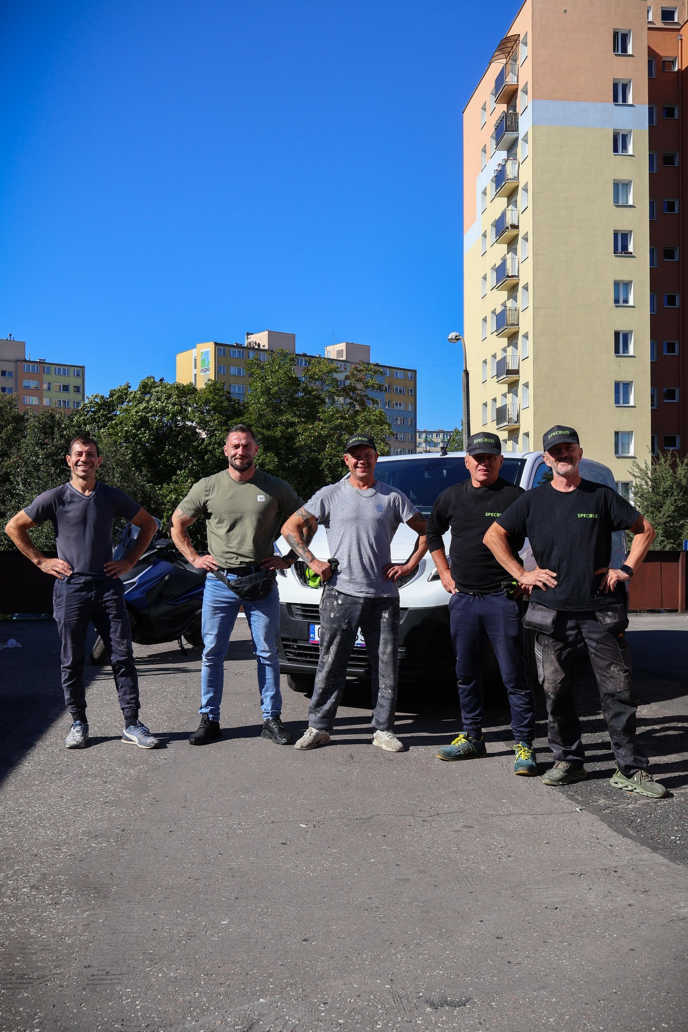 Zespół pracowników budowlanych w firmowych koszulkach i czapkach, stojący przed budynkiem mieszkalnym i samochodem dostawczym w słoneczny dzień.