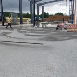 Prace brukarskie przy nowej hali: ułożona szara kostka, pracownicy z taczką, widoczny drenaż liniowy i przygotowane miejsce na instalacje. W tle sprzęt budowlany.