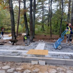 Zakład Remontowo Budowlany Michał Frątczak - Układanie kostki brukowej w parku: pracownik z wózkiem, szare i białe kostki z żółtymi elementami dla niewidomych, drzewa w tle, jesienna sceneria.