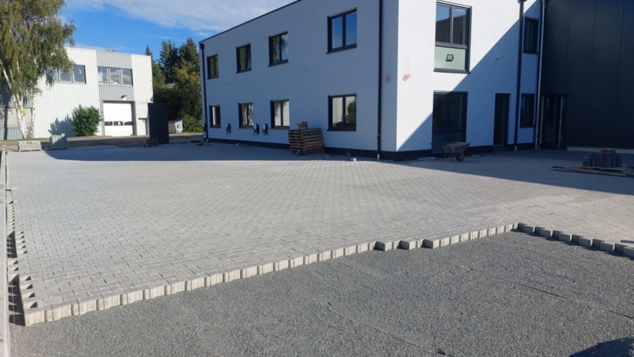 Szara kostka brukowa ułożona na posesji przy budynku. Widoczne obrzeża i przygotowane podłoże. W tle budynki i drzewa.