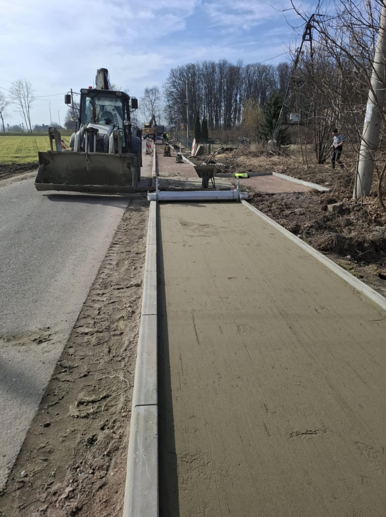 Przygotowanie podłoża pod kostkę: równanie piasku z użyciem listwy wibracyjnej, z tyłu koparko-ładowarka i pracownicy. Etap prac przed ułożeniem kostki brukowej.