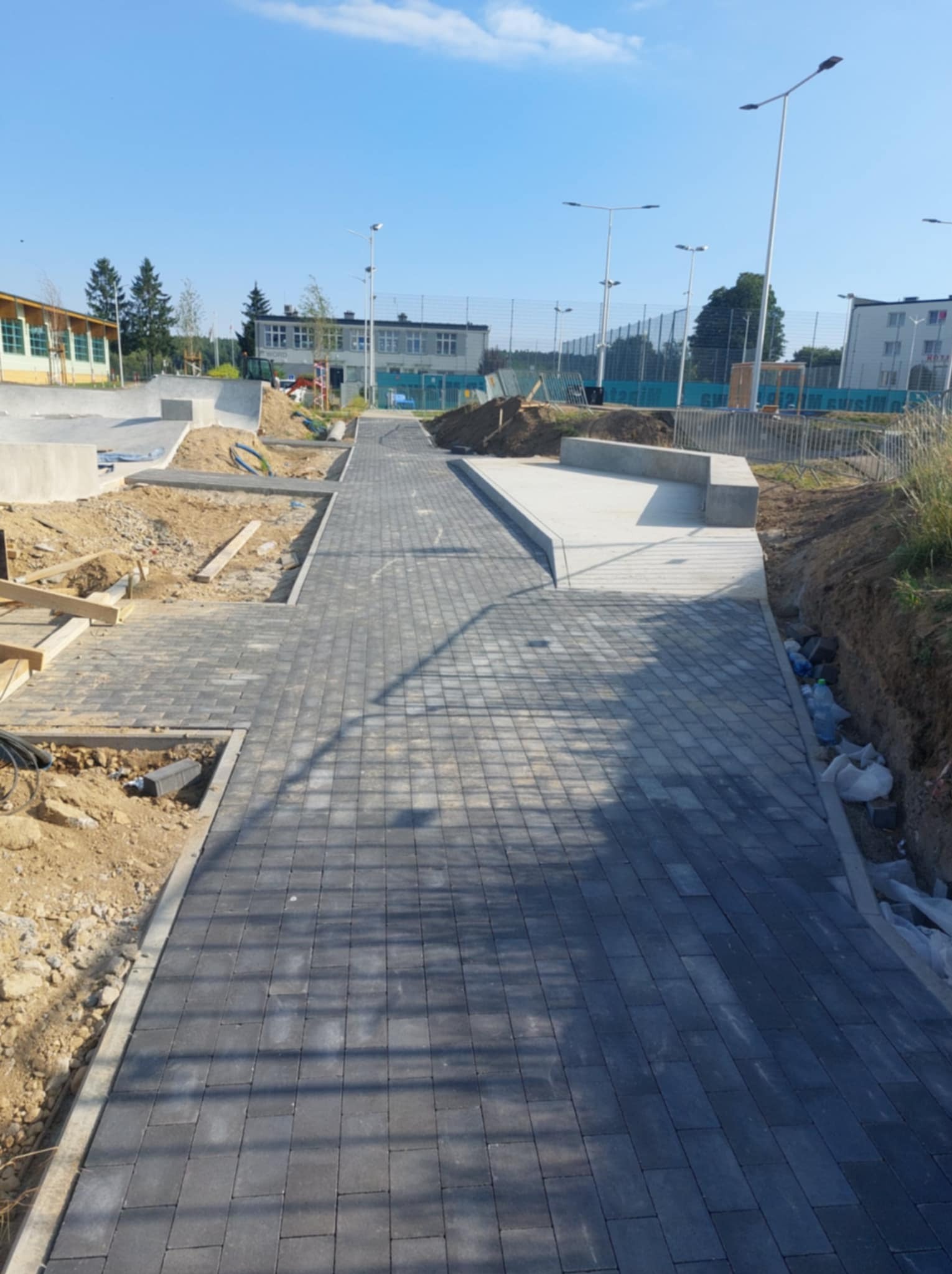 Ścieżka z szarej kostki brukowej w trakcie budowy, z widocznymi elementami skateparku i oświetleniem ulicznym w tle, w słoneczny dzień.