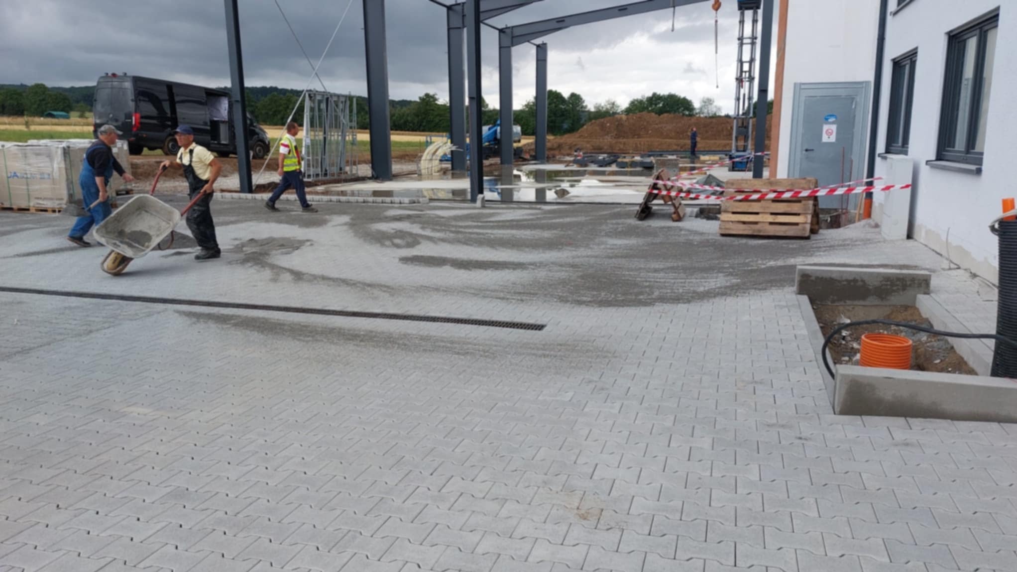 Prace brukarskie przy nowej hali: ułożona szara kostka, pracownicy z taczką, widoczny drenaż liniowy i przygotowane miejsce na instalacje. W tle sprzęt budowlany.