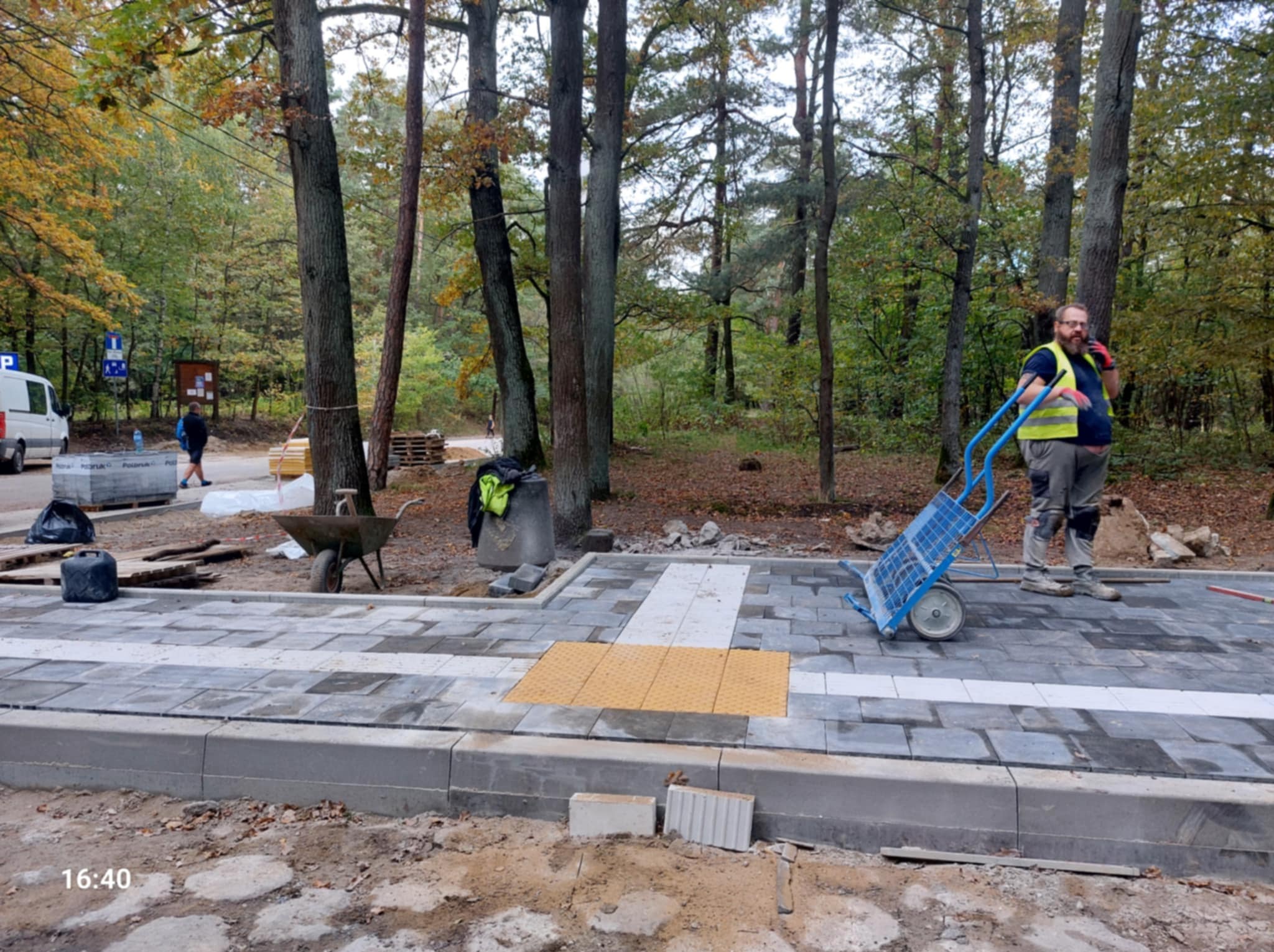 Układanie kostki brukowej w parku: pracownik z wózkiem, szare i białe kostki z żółtymi elementami dla niewidomych, drzewa w tle, jesienna sceneria.