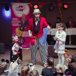 Iluzjonista w czerwonym fraku i pasiastej spodniach wykonuje trik dla dzieci przebranych w kostiumy na tle sceny z plakatem cyrkowym.