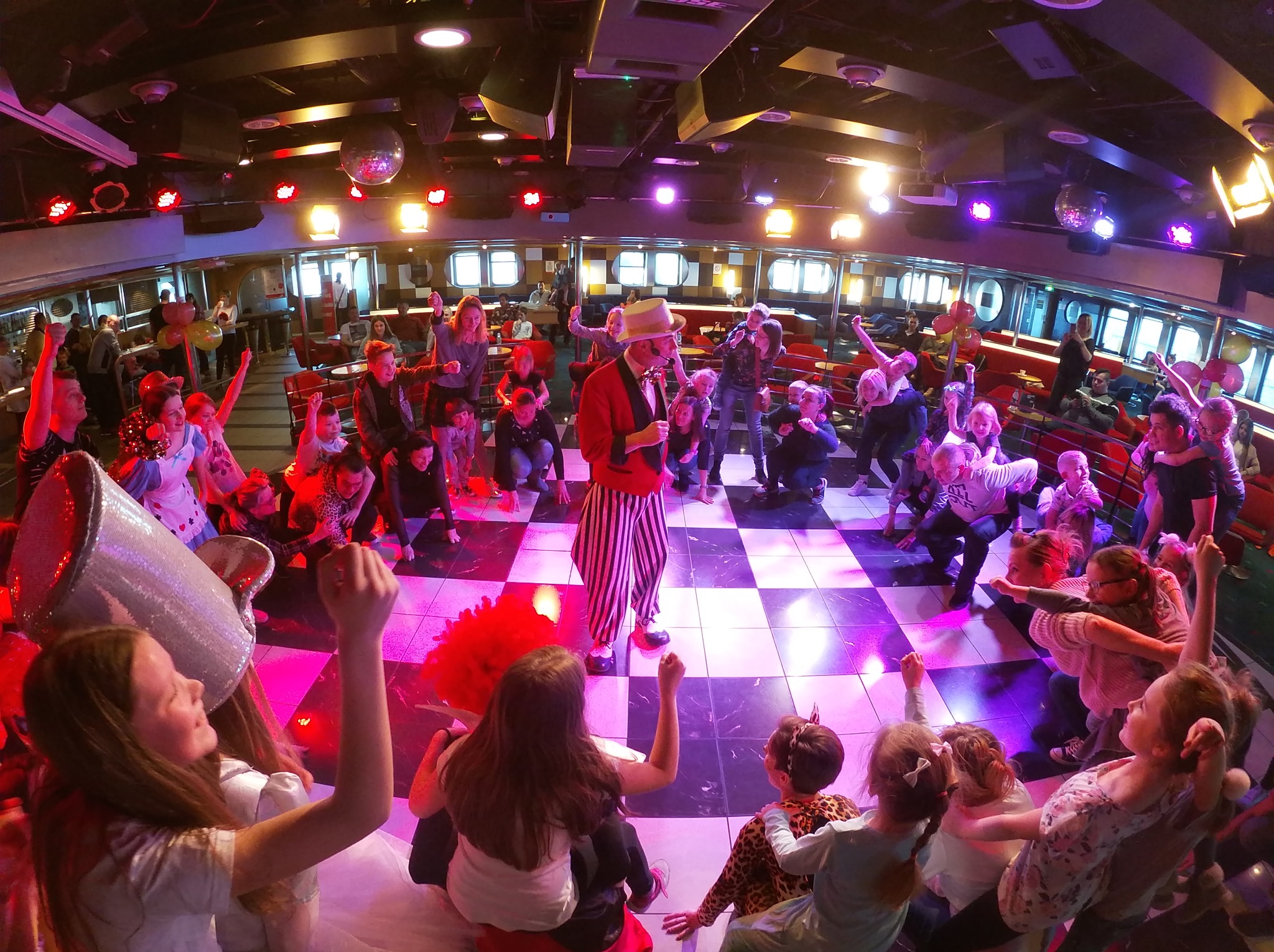 Animator w czerwonym fraku i paski prowadzi zabawę dla dzieci na statku, oświetlonym kolorowymi światłami i dyskotekową kulą. Dynamiczna scena z uśmiechniętymi uczestnikami.