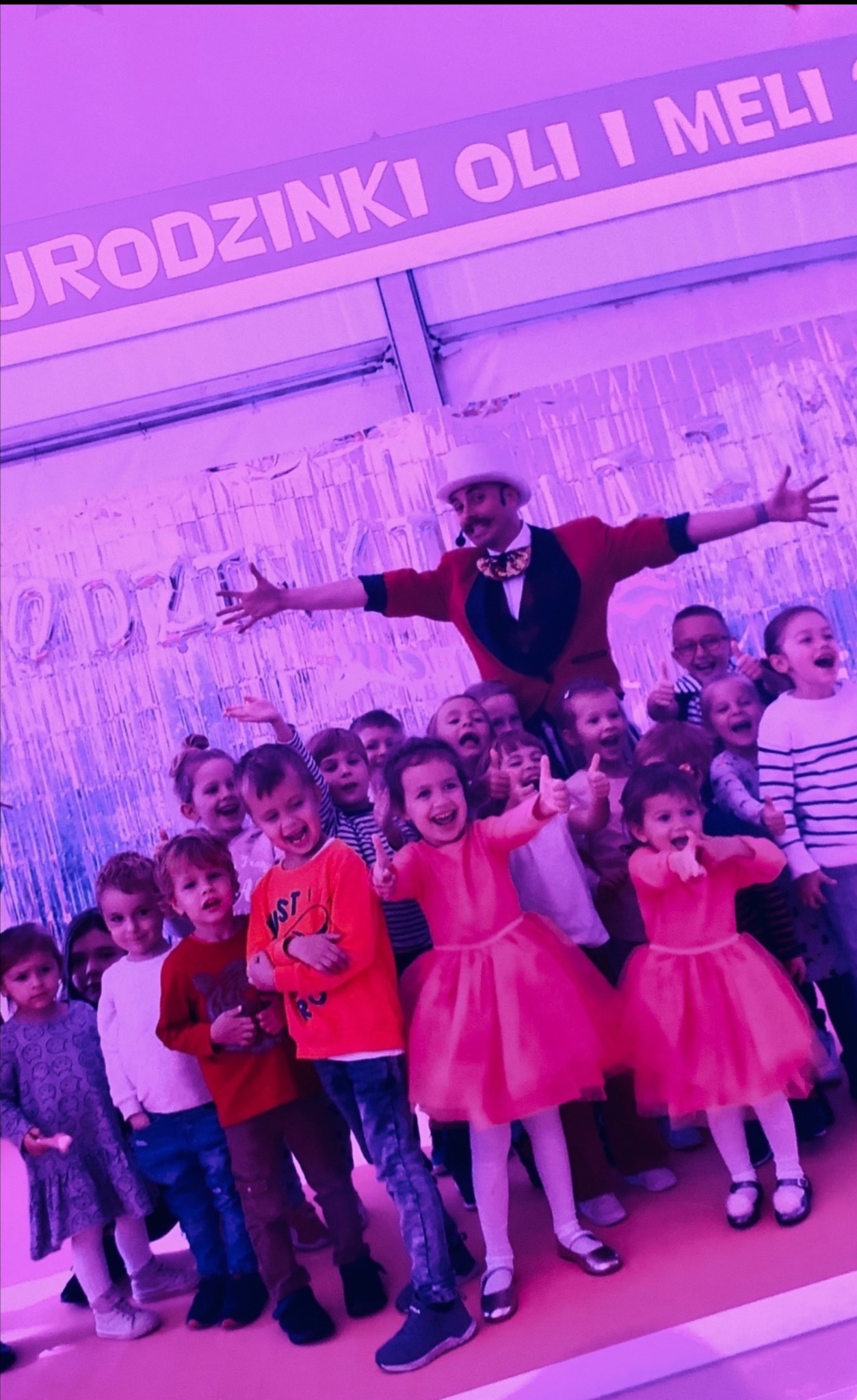 Uśmiechnięty iluzjonista w cylindrze i czerwonej marynarce otoczony grupą dzieci na tle dekoracji urodzinowych w odcieniach fioletu.