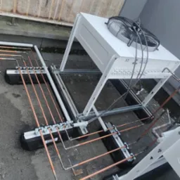 Montaż instalacji chłodniczej na dachu budynku. Widoczne miedziane rury, wsporniki i jednostka zewnętrzna klimatyzacji z wentylatorem, zamontowana na konstrukcji wsporczej.