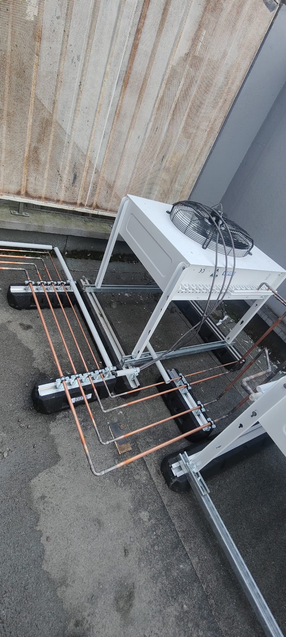 Montaż instalacji chłodniczej na dachu budynku. Widoczne miedziane rury, wsporniki i jednostka zewnętrzna klimatyzacji z wentylatorem, zamontowana na konstrukcji wsporczej.