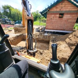 Adrian Czepiela - Z wnętrza kabiny koparki widok na wykop pod budynek z cegły. W tle zieleń i fragment placu budowy. Widoczne ramię koparki i elementy sterujące.