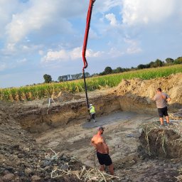 Adrian Czepiela - Wylewanie betonu do wykopu pod fundamenty z użyciem pompy. Widoczni pracownicy w trakcie pracy, teren budowy otoczony polem uprawnym, błękitne niebo z chmurami.