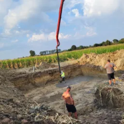 Wylewanie betonu do wykopu pod fundamenty z użyciem pompy. Widoczni pracownicy w trakcie pracy, teren budowy otoczony polem uprawnym, błękitne niebo z chmurami.
