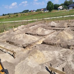 Adrian Czepiela - Wykopane rowy pod fundamenty domu jednorodzinnego z drewnianymi znacznikami, widok z kabiny maszyny budowlanej na tle zieleni i błękitnego nieba.