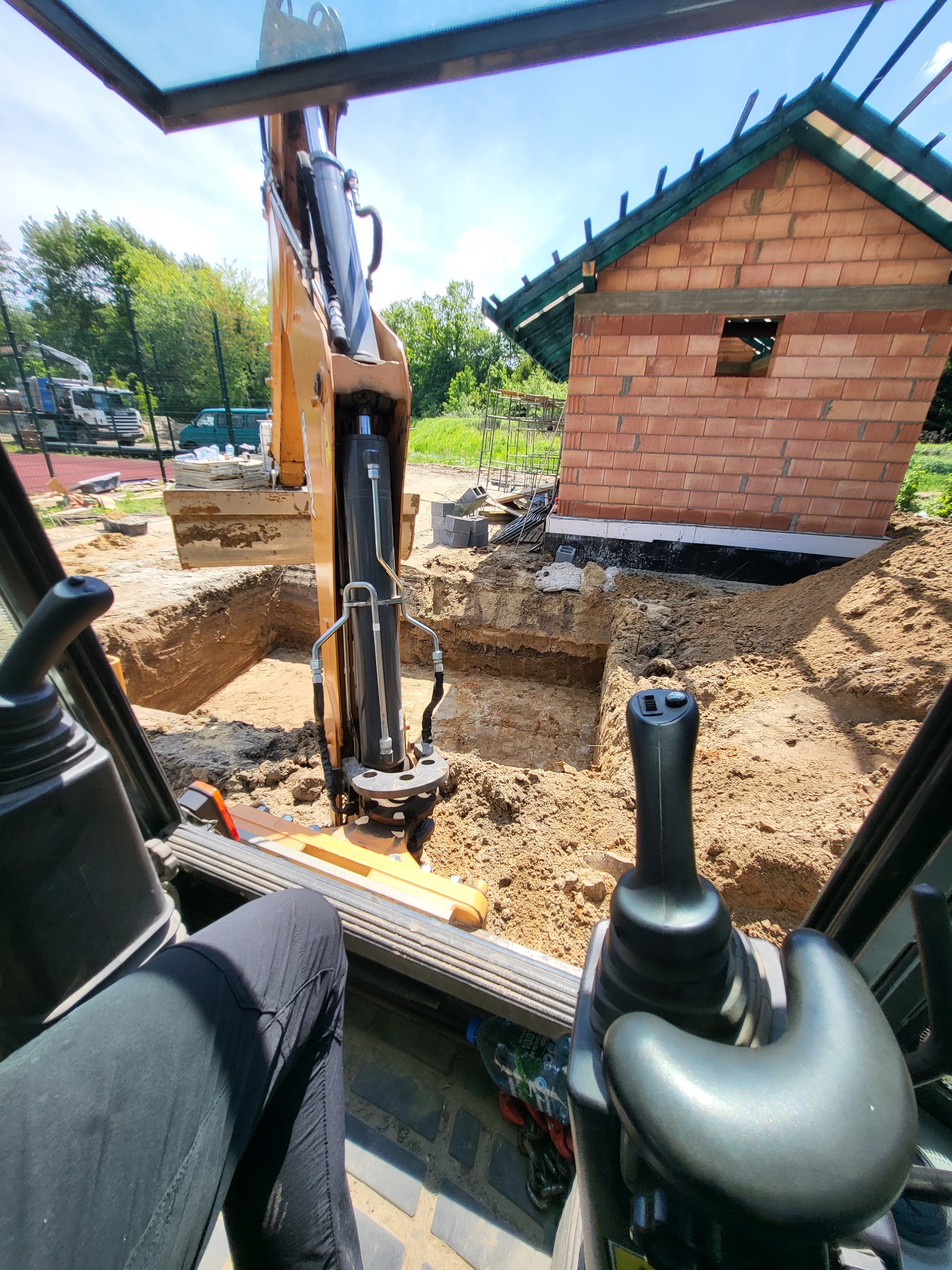 Z wnętrza kabiny koparki widok na wykop pod budynek z cegły. W tle zieleń i fragment placu budowy. Widoczne ramię koparki i elementy sterujące.