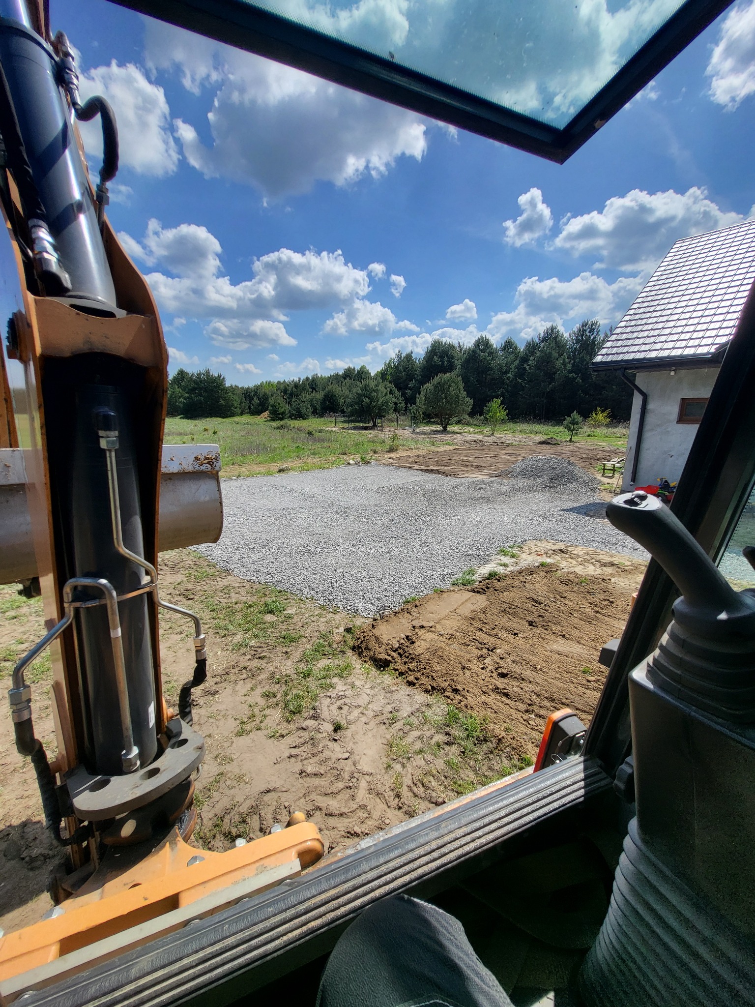 Widok z kabiny koparki na utwardzony teren żwirem przy domu jednorodzinnym, w tle las i błękitne niebo z chmurami. Prace ziemne w toku.
