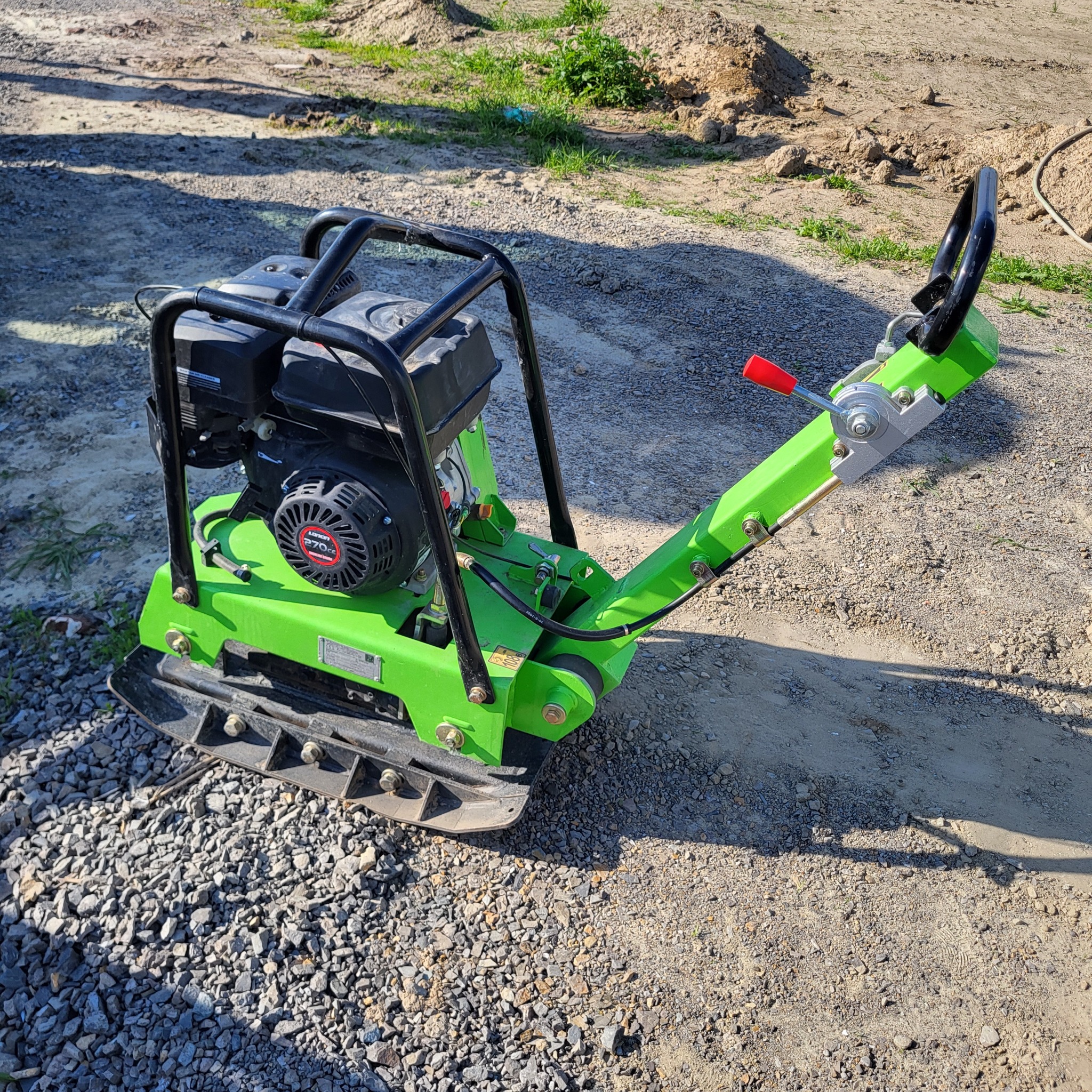 Zielona zagęszczarka spalinowa na żwirze, w tle ziemia. Sprzęt budowlany do utwardzania terenu, widoczne ślady użytkowania. Słoneczny dzień.