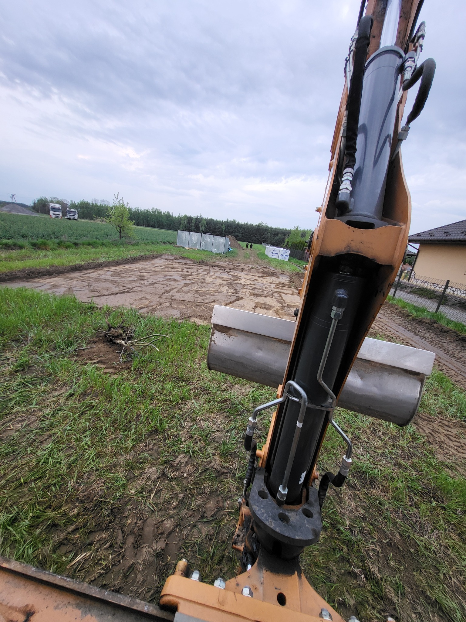 Widok z perspektywy operatora na ramię koparki i przygotowany teren pod budowę, z widocznymi śladami opon i ogrodzeniem w tle.