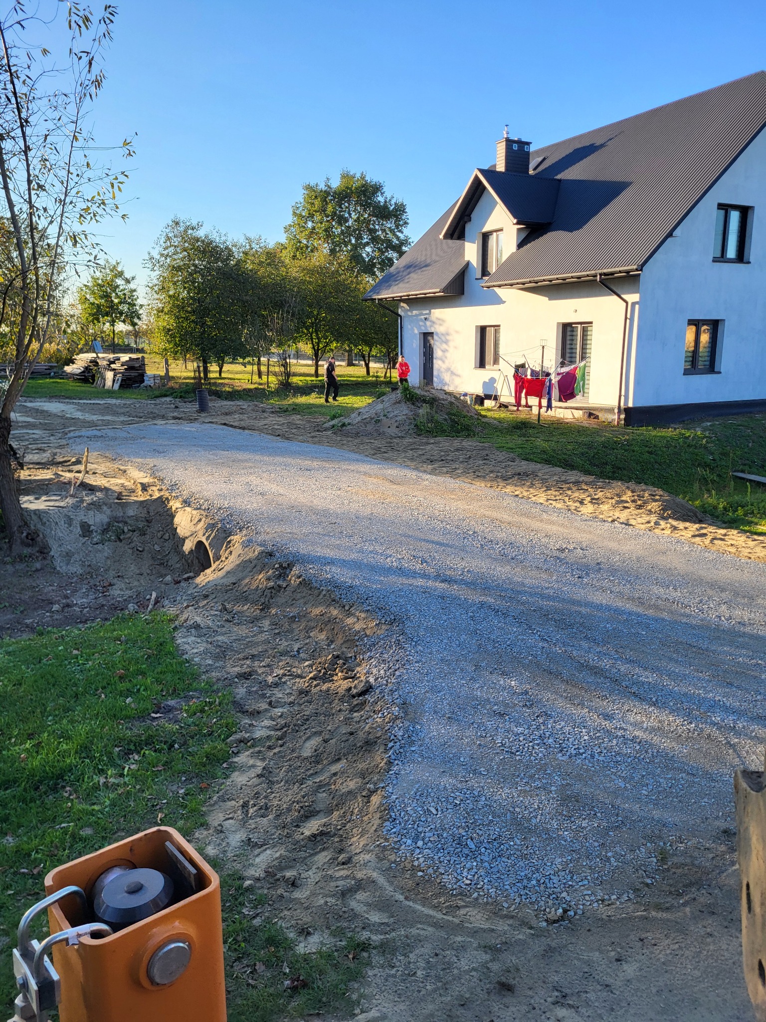 Utwardzona żwirowa droga do domu z widocznym przepustem pod nią. W tle dom z szarym dachem, drzewa i osoba. Jasne niebo.