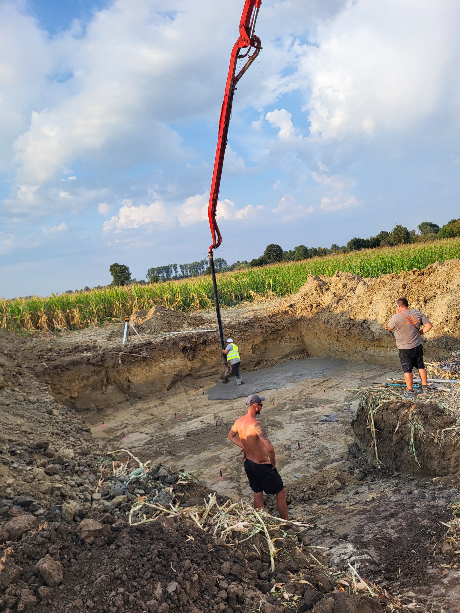 Wylewanie betonu do wykopu pod fundamenty z użyciem pompy. Widoczni pracownicy w trakcie pracy, teren budowy otoczony polem uprawnym, błękitne niebo z chmurami.