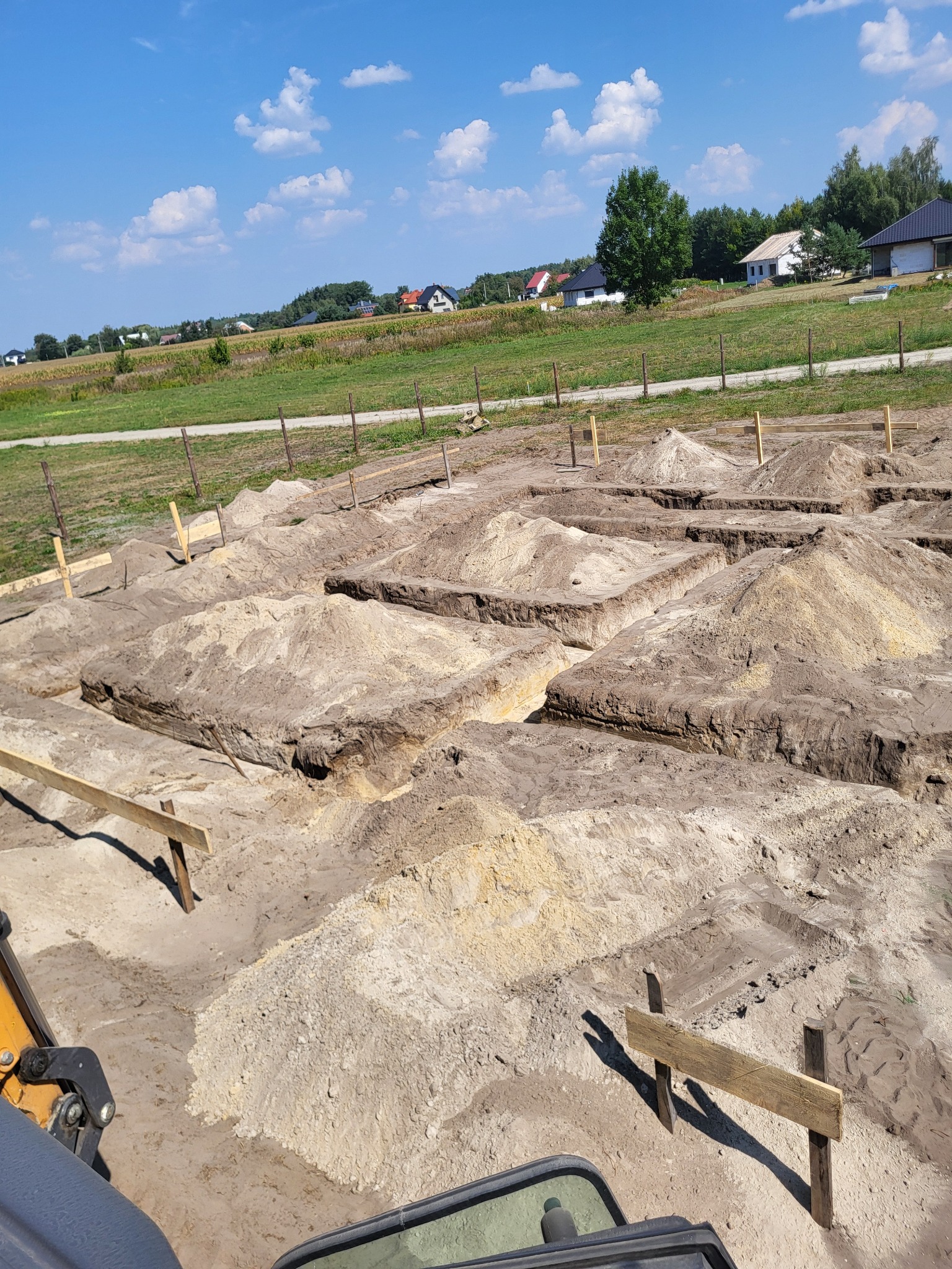 Wykopane rowy pod fundamenty domu jednorodzinnego z drewnianymi znacznikami, widok z kabiny maszyny budowlanej na tle zieleni i błękitnego nieba.