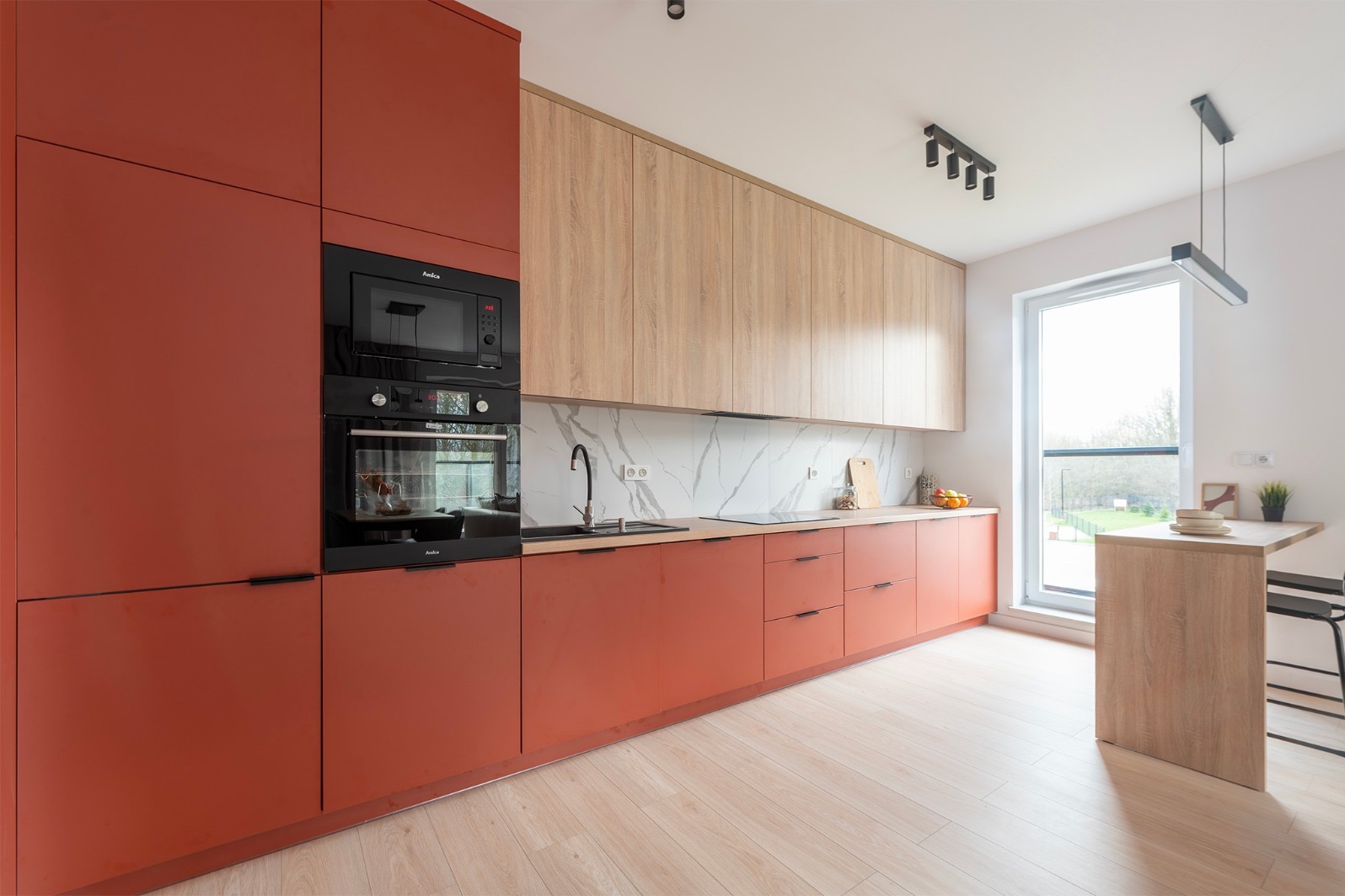 Nowoczesna kuchnia z terakotowymi szafkami, drewnianymi górnymi szafkami i marmurowym backsplashiem. Wbudowane urządzenia Amica. Jasna podłoga i stół barowy przy oknie.