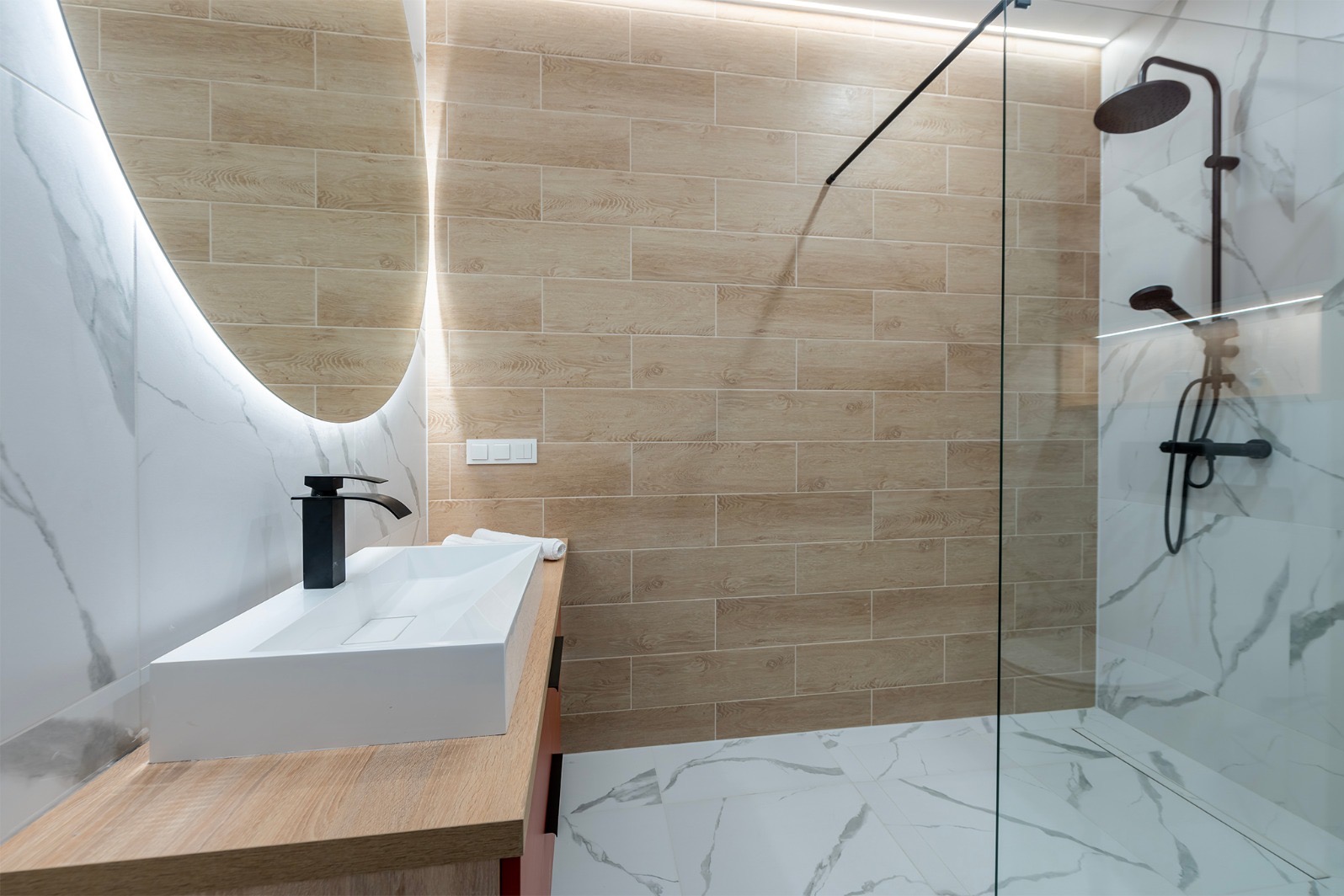Nowoczesna łazienka z beżowymi płytkami imitującymi drewno, białą umywalką nablatową i okrągłym lustrem z podświetleniem LED. Kabina prysznicowa ze szklaną ścianką.