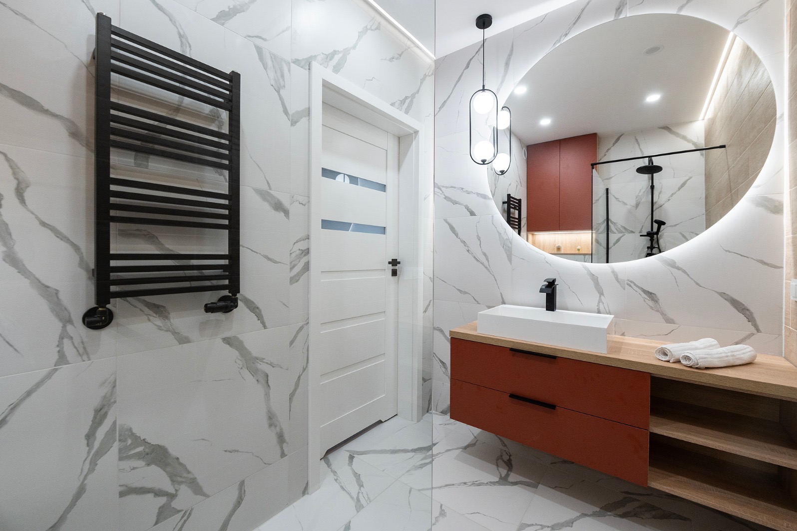 Nowoczesna łazienka z marmurowymi płytkami, okrągłym lustrem z podświetleniem LED, wiszącą szafką w kolorze terakoty i czarnym grzejnikiem drabinkowym.