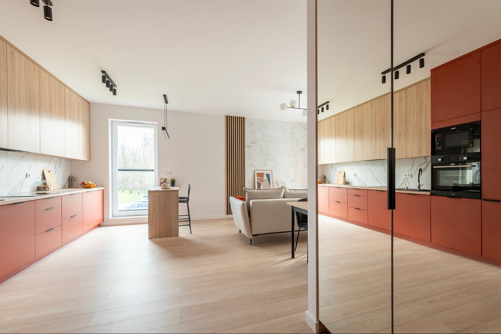 Nowoczesna kuchnia z salonem: drewniane i koralowe szafki, marmurowa ściana, lustrzane drzwi, parkiet. Widok na salon z sofą i jadalnią. Oświetlenie punktowe.