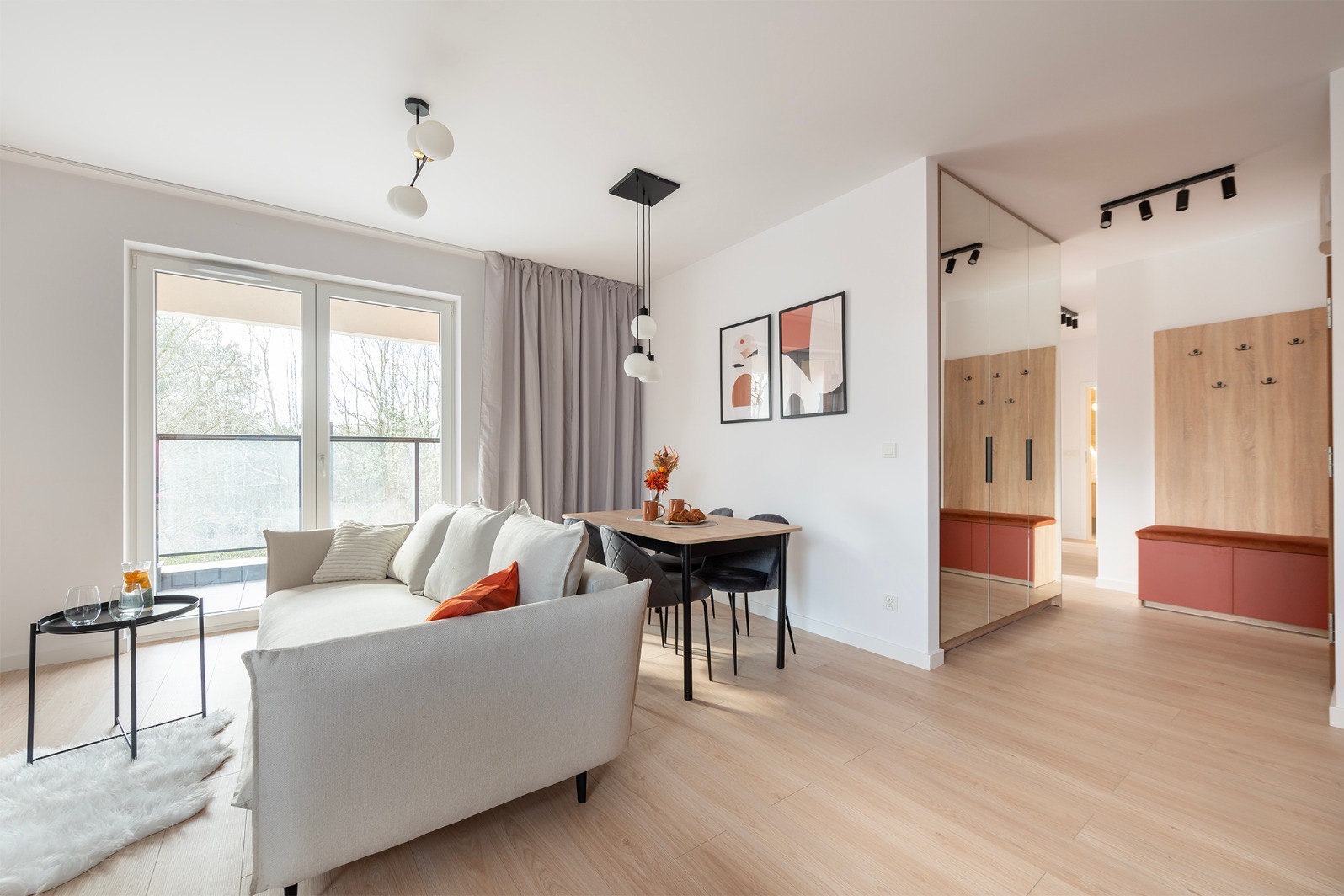 Jasne wnętrze mieszkania z sofą, stołem jadalnym i przedpokojem z lustrem. Minimalistyczny design, drewniana podłoga, naturalne światło. Nowoczesne oświetlenie.