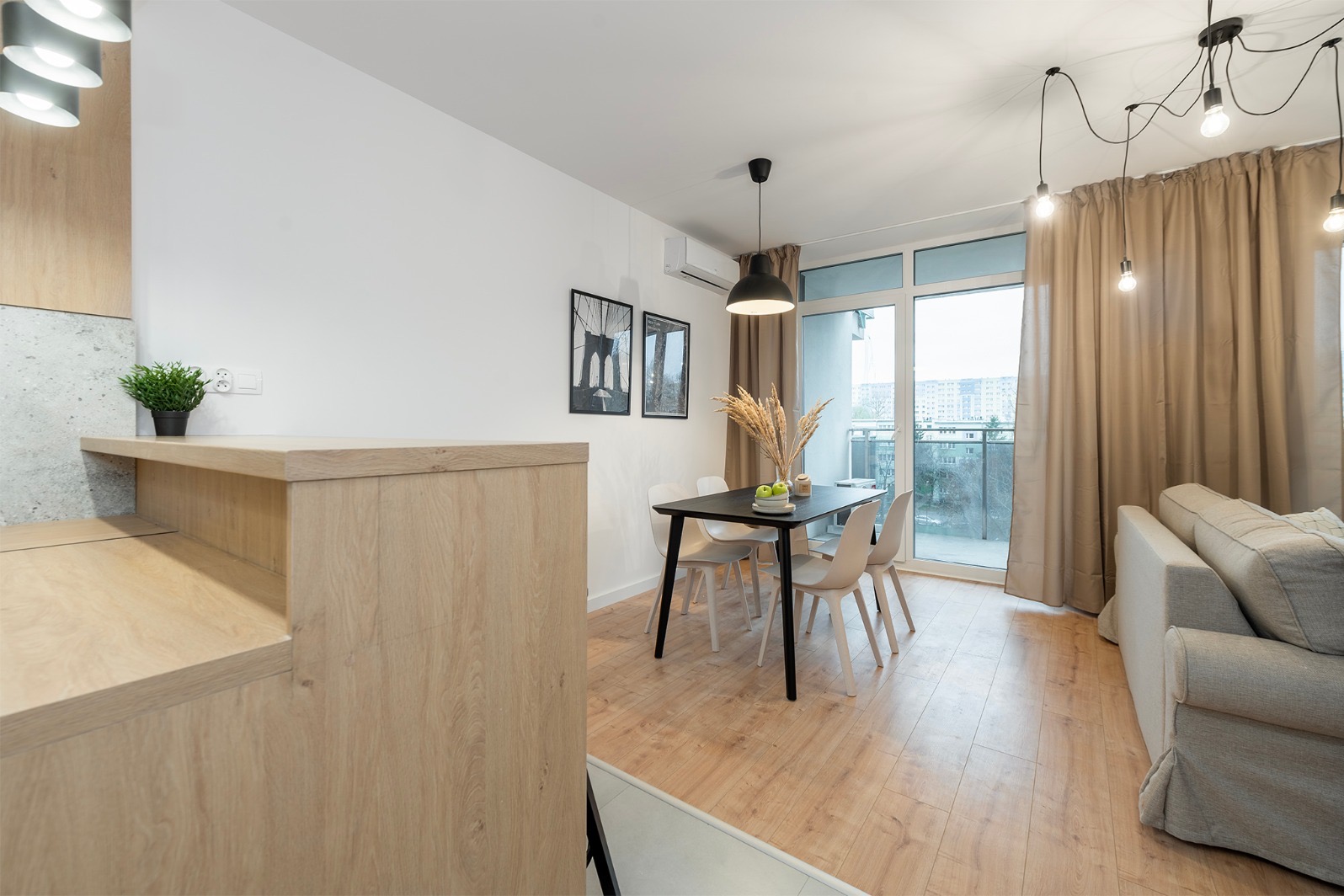 Jasne wnętrze: stół z krzesłami, sofa pod oknem balkonowym z beżowymi zasłonami. Minimalistyczne lampy wiszące. Drewniana podłoga i elementy wykończenia.