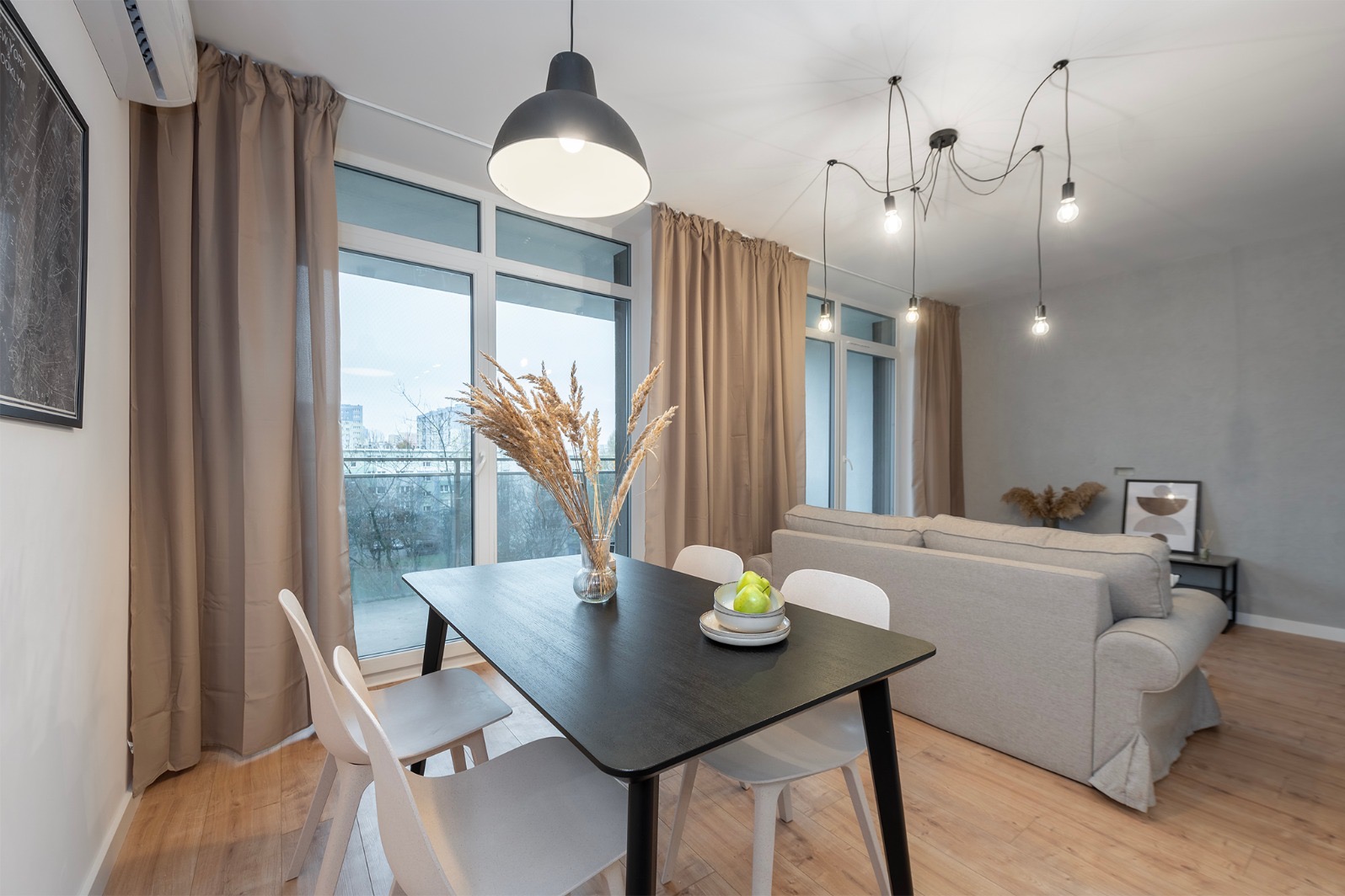 Jasne wnętrze: czarny stół z dekoracją, białe krzesła, beżowa sofa, okno balkonowe z zasłonami. Nowoczesne lampy wiszące. Minimalistyczny design.